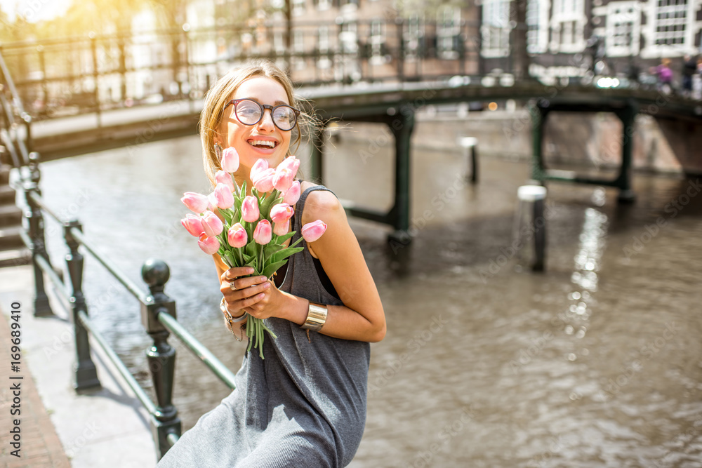 Fototapeta premium Młoda kobieta zabawny siedzi z bukietem różowych tulipanów w pobliżu kanału wodnego w mieście Amsterdam