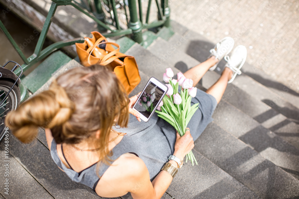 Fototapeta premium Young woman photographing with phone a bouquet of pink tulips sitting outdoors in Amsterdam city