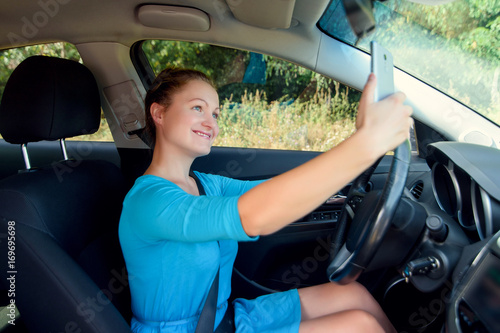 Happy young woman holding mobile phone and taking photos while driving a car. Smiling girl taking selfie picture with smart phone camera outdoors in car. Holidays and tourism concept