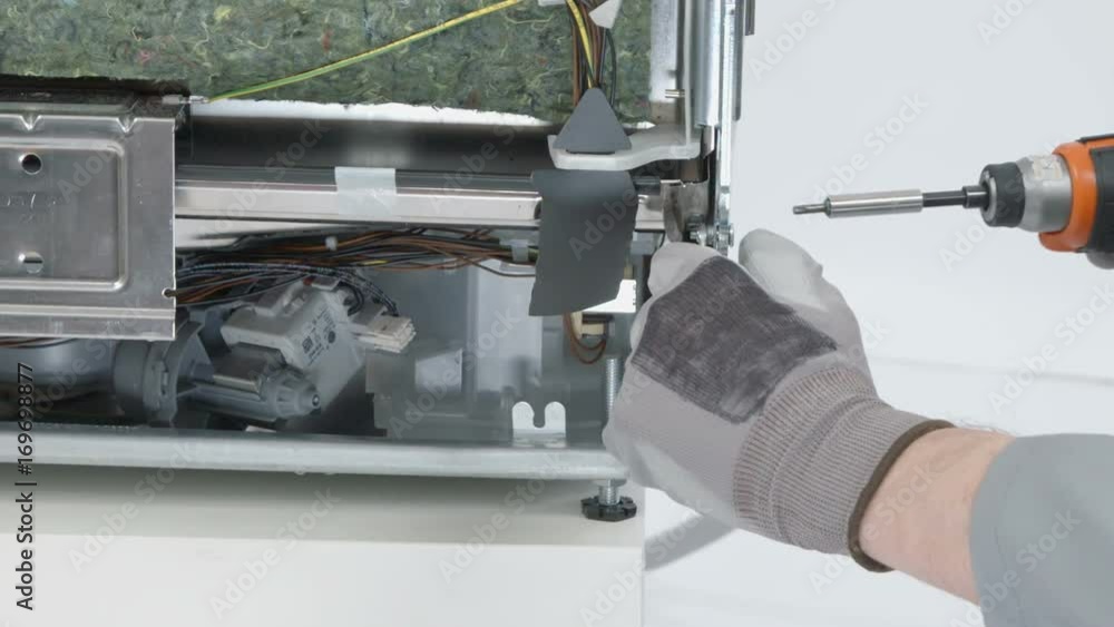 A young repairman is using a drill in order to take out a nail on the bottom of the machine. He is wearing safety clothes and gloves. A young repairman is using a drill in order to take out a nail on the bottom of the machine. He is wearing safety clothes and gloves.