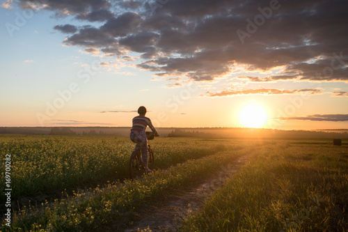 Girl on a bicycle in a field on the road against the sunset background © Sergei Malkov