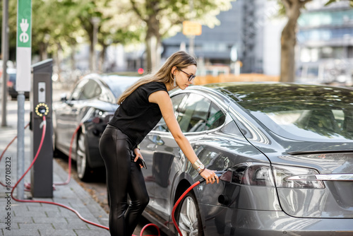 Young woman in black clothes putting connector into the electric car outdoors on the street in Rotterdam city