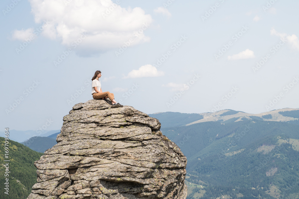 Young woman with wind in her hair sits on a rock  in mountain rural area on sunny summer day and enjoys beautiful scenery. Person in casual clothing poses on a cliff and looks into far away distance