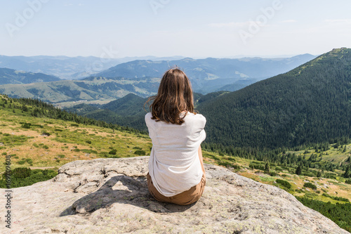 Back of young female person sitting on top of the rock in ukrainian carpathian mountains. Girl tourist enjoys gorgeous landscape scenery on hot sunny day