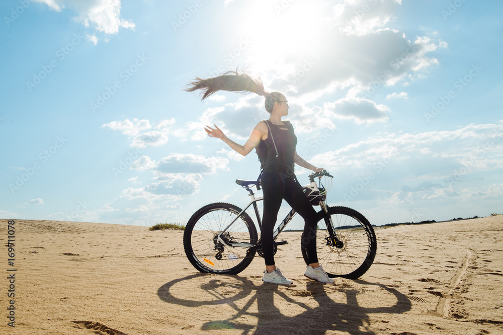 Fototapeta premium Woman cyclist carrying mountain bike on sunrise trail. Girl in sport wear on black white bicycle going to record in summer sunset time in desert.