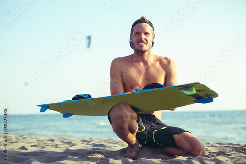 Smiling Caucasian man kitesurfer sittingg on the beach with his board
