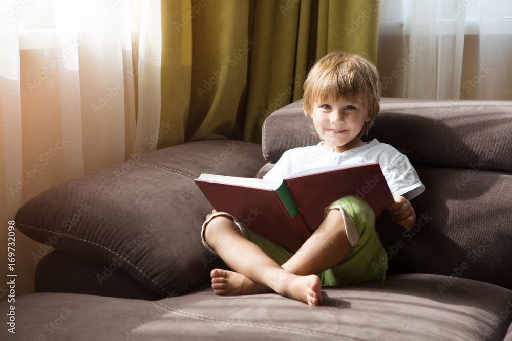 Cute little boy is reading book sitting on a sofa at home Cute little boy is reading book sitting on a sofa at home