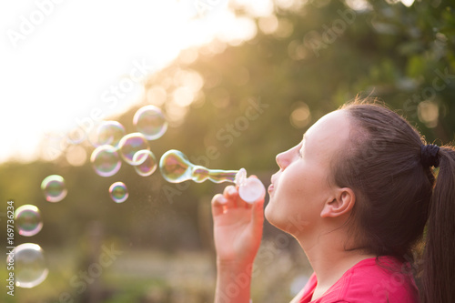 Beautiful young brunette girl blowing soap bubbles outdoor during lovely summer sunset