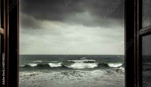 Dramatic sea view from opened window with big stormy waves and dramatic sky during rain and storm weather in fall season on sea coast