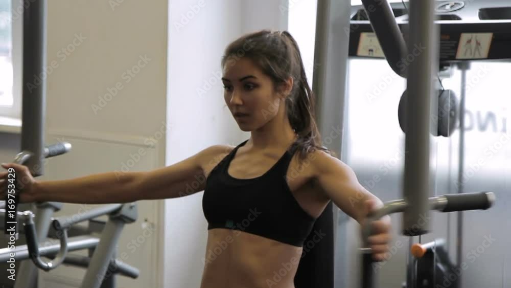 Young woman doing exercise on pec deck machine in gym indoors. Female chooses working weight, sits on bench, holds on to handles, with exhalation, spreads hands wide apart and returns to starting Young woman doing exercise on pec deck machine in gym indoors. Female chooses working weight, sits on bench, holds on to handles, with exhalation, spreads hands wide apart and returns to starting