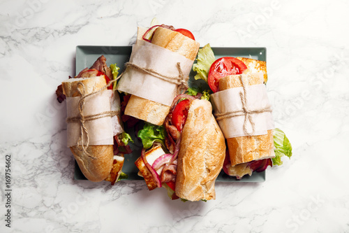 Fresh baguette sandwich bahn-mi styled. Bacon, roasted cheese, tomatoes and lettuce on metallic tray on white marble background.