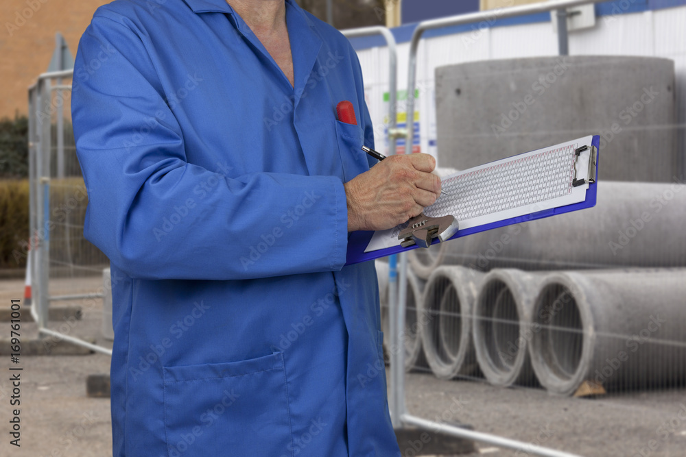 Plumber in blue overalls checking a data sheet on clipboard while holding a spanner and standing in front of a pile of concrete drainage pipes on a construction site Plumber in blue overalls checking a data sheet on clipboard while holding a spanner and standing in front of a pile of concrete drainage pipes on a construction site