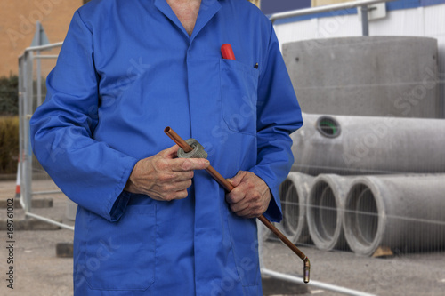 Plumber in blue overalls cutting a copper pipe while standing in front of a pile of concrete drainage pipes on a construction site