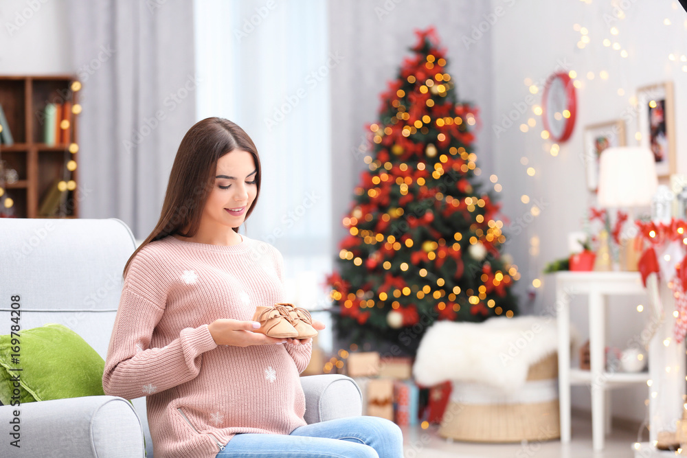 Happy pregnant woman with baby booties in room decorated for Christmas Happy pregnant woman with baby booties in room decorated for Christmas