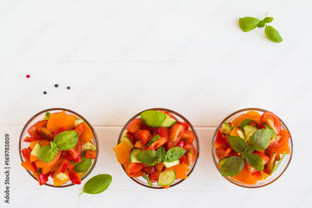 Vegetarian vegetable salad with tomatoes, cucumber, red bell pepper and basil in the glass bowls on the white wooden table, top view. Vegetarian vegetable salad with tomatoes, cucumber, red bell pepper and basil in the glass bowls on the white wooden table, top view.