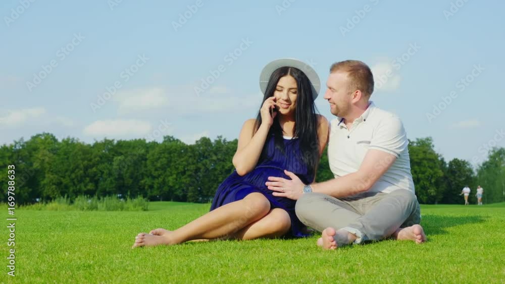 Young pregnant woman speaks by phone. Next to her sits her husband, together rest in the park Young pregnant woman speaks by phone. Next to her sits her husband, together rest in the park