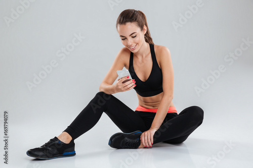 Smiling fitness woman sitting on the floor and using smartphone