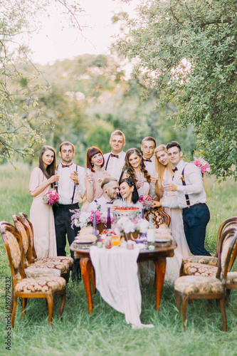 The vertical photo of the happy newlywed couple and the smiling guests standing behind them at the background of the spring green field. photo