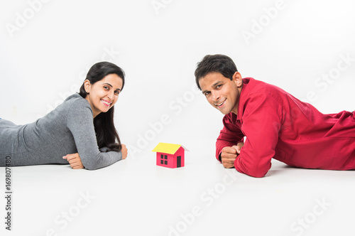 Canvas Print stock photo showing side angle of Indian or asian smart and cheerful / happy couple holding 3D paper house model and sitting isolated over white background, asian couple and real estate concept