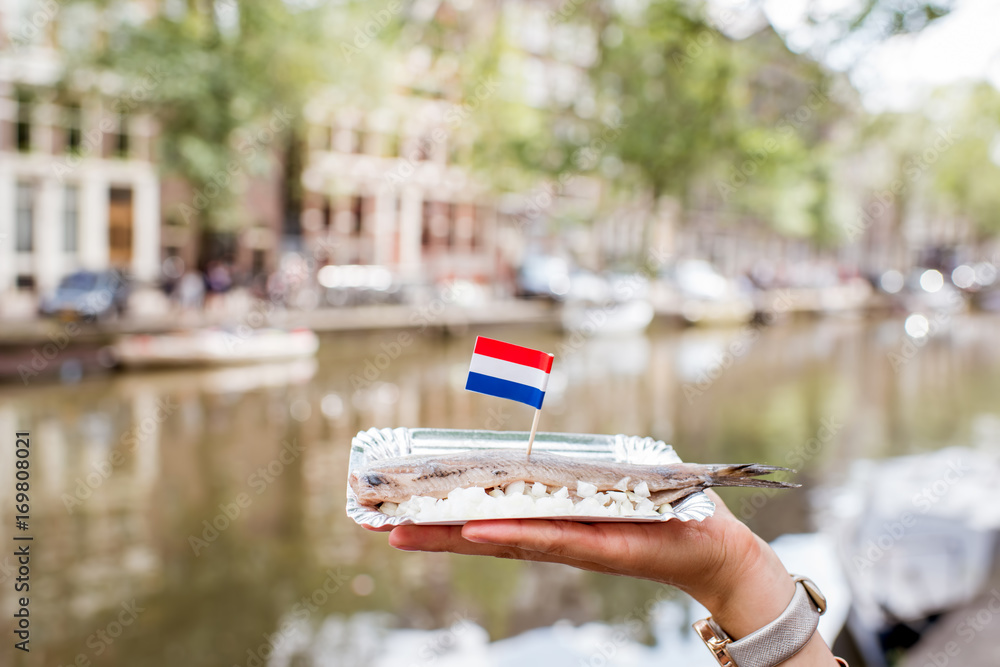 Naklejka premium Trzyma świeży harring z cebulą i flagą Holandii na tle kanału wodnego w Amsterdamie