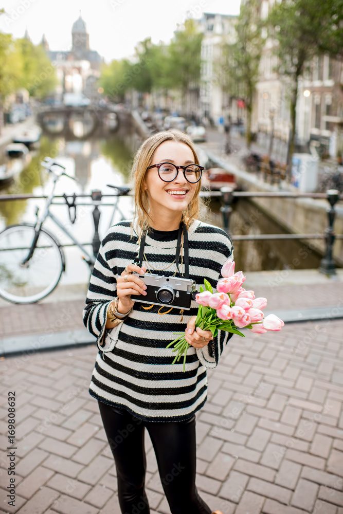 Fototapeta premium Portrait of a young woman tourist with photo camera and flowers standing on the bridge in Amsterdam old city