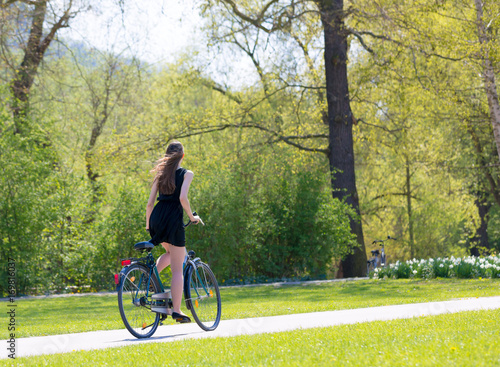 Portrait view of girl on bicycle wearing on black short dress. Young happy Woman riding along road on green spring  outdoor Park. Sporty young girl riding a bicycle on a sunny morning