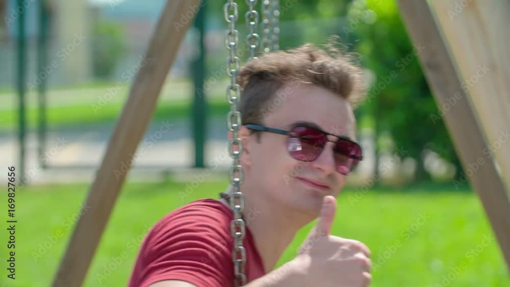 A teenager is swinging on the swing. It is a nice and sunny day and he would like to relax a bit.