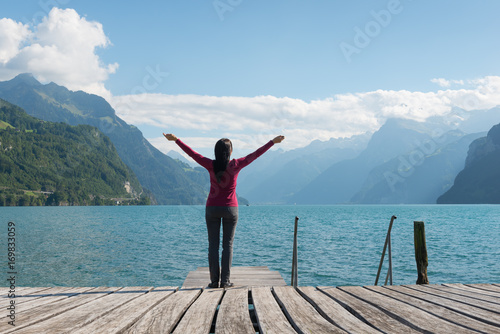 Woman with open arms standing on the shore of the lake. Mountain range in the background.