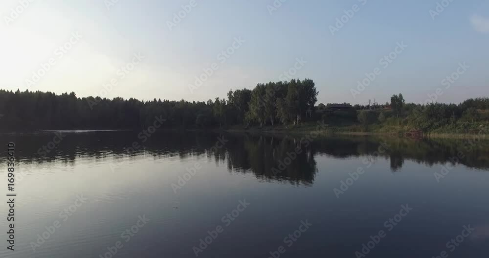 Moscow oblast, Russia. Camera flight over the lake, clear like a mirror. It reflects the trees on the shore, green grass, wooden house and blue sky. Moscow oblast, Russia. Camera flight over the lake, clear like a mirror. It reflects the trees on the shore, green grass, wooden house and blue sky.