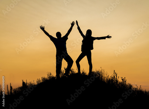 Two people between clouds Full length silhouette of happy couple stand together on peak of a mountain with hands raised up  sunset and ocean. Man and woman on top mountain look at beautiful night