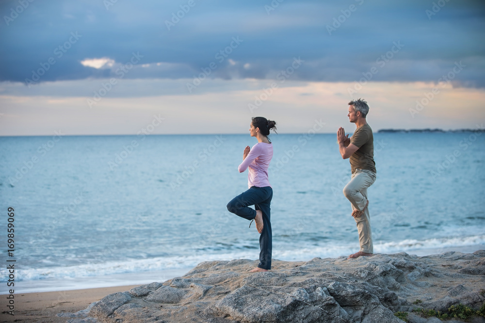 Beautiful couple practicing yoga on the beach at sunrise
