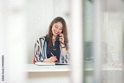 Cheerful young businesswoman is talking on the phone while working at her desk