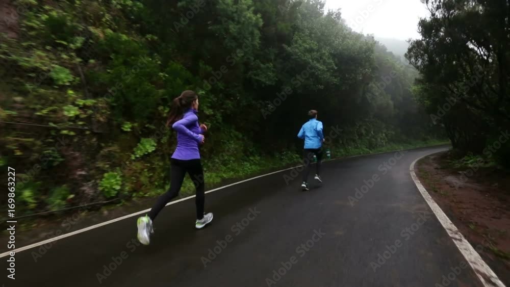 Fitness cport couple jogging. Tracking shot of man and woman are running on wet country road. Determined couple are jogging by mountains. Sporty people are representing their healthy lifestyle. Fitness cport couple jogging. Tracking shot of man and woman are running on wet country road. Determined couple are jogging by mountains. Sporty people are representing their healthy lifestyle.