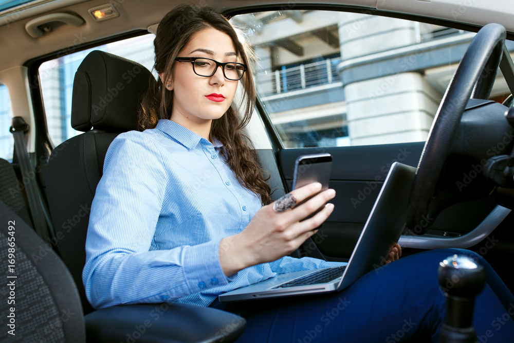 Beautiful young business woman sitting in the car with laptop looking at her smart phone.