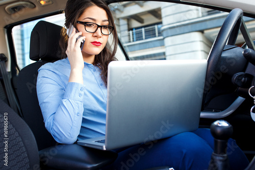 Beautiful young business woman sitting in the car with laptop and talking on phone. © Zoran Zeremski