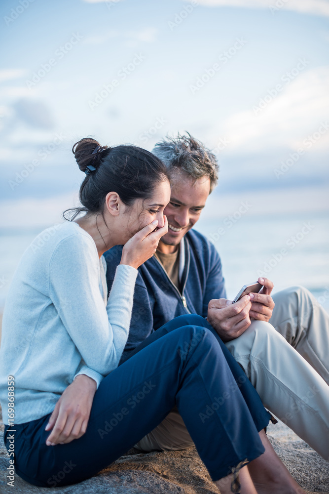 A couple sitting at the beach, looking at photos on a phone