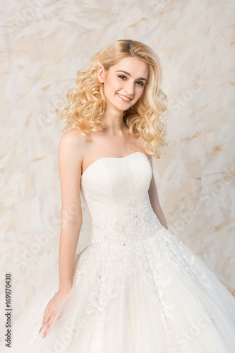 fashionable white gown, beautiful blonde model, bride hairstyle and makeup concept - young smiling girl in wedding festive dress, standing indoors on light background, romantic slender woman posing