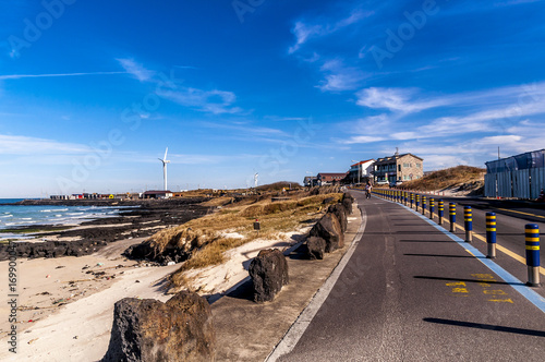Beautiful view Woljeong Beach besides trunk road providing leading line at Jeju Island South Korea with white windmill at far left backed by awesome blue sky background