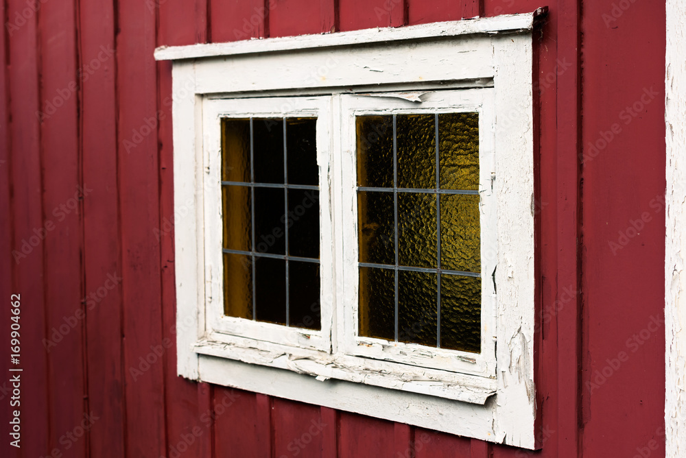 Old window on a red cottage with white window frame. The white frame is flaking and in need of attention. Lead mullions or glazing bars on the glass window.