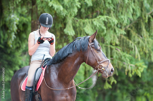 Chestnut horse with teenage girl with smartphone sitting on it