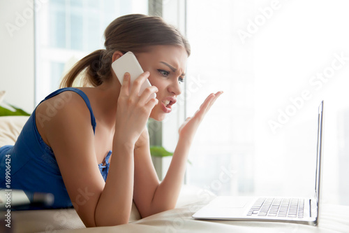 Angry woman lying on sofa in front of laptop and arguing on cellphone. Jealous lady calling her boyfriend because of photos online, explaining problem to support service, annoyed by work on weekend