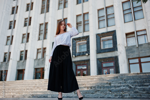 Portrait of a fabulous young successful woman in white blouse and broad black pants posing on the stairs with a huge white building on the background.