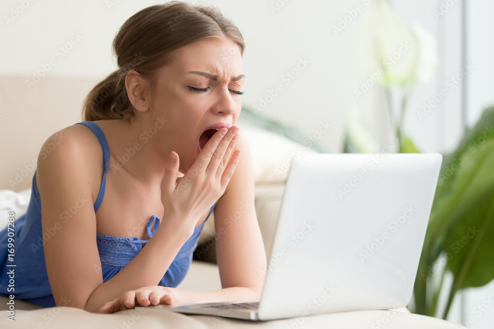 Bored young woman lying on sofa in front of laptop and yawning. Attractive lady boring because of uninteresting movie, tired after too long work on computer, feeling lack of sleep when resting at home Bored young woman lying on sofa in front of laptop and yawning. Attractive lady boring because of uninteresting movie, tired after too long work on computer, feeling lack of sleep when resting at home