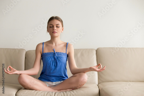 Charming young lady in casual clothes meditating on couch in lotus position, doing yoga exercises in living room in morning, relaxing and making autosuggestion practices to get positive thinking photo