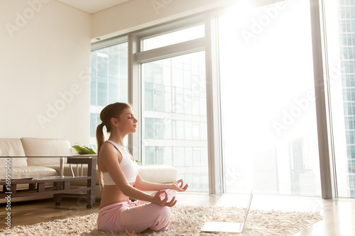 Attractive young woman wearing comfortable sportswear relaxing, meditating on living room carpet. Calm lady sitting on floor in lotus position in front of laptop, listening spiritual practices lessons photo