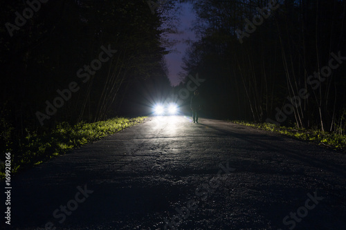Male silhouette at the edge of a dark mountain road through the forest in the night. Man standing on the road against the car headlights. The car on the roadside. Mystery concept. Soft focus, filter.