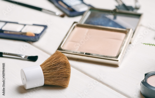 Cosmetic brush with open powder box among other cosmetics on a white wooden table