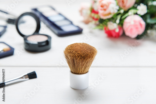 Big cosmetic brush among makeup items on a white wooden table with roses bouquet on the background, closeup shot, selective focus