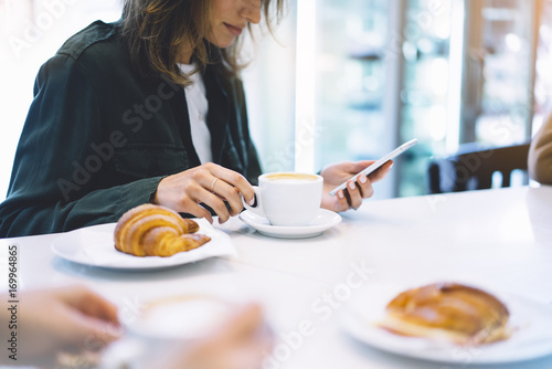 Young woman holding in female hands mobile phone and drink hot aroma coffee or tea in breakfast time, hipster traveler girl in the morning using smartphone; holiday relax white cafe