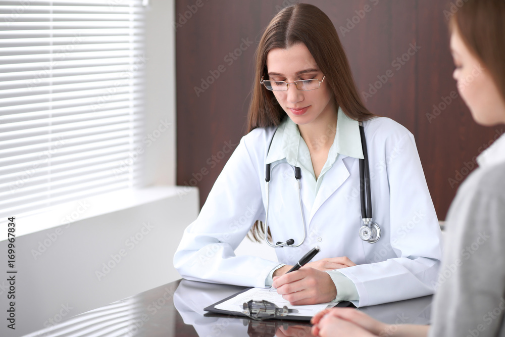 Female doctor having a talk with her patient while sitting at the table near the window in hospital. Physician is ready to help patient. Medicine and health care concept Female doctor having a talk with her patient while sitting at the table near the window in hospital. Physician is ready to help patient. Medicine and health care concept